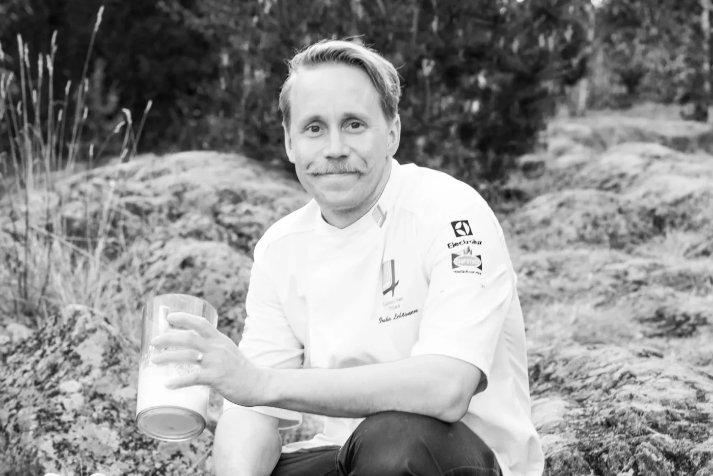Juha Lehtonen hymyilee luonnon keskellä, yllään Suomen kokkimaajoukkueen kokkitakki.